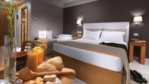 Une chambre d’hôtel avec un grand lit, des tables de chevet, une petite table, un vase avec des fleurs, un plateau de pâtisseries, une théière et un verre de jus d’orange.