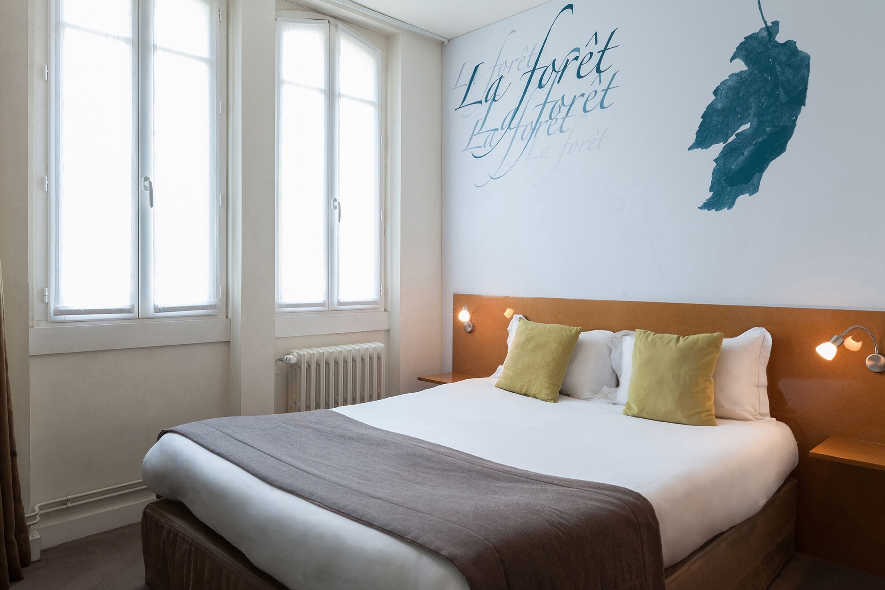 Une chambre d’hôtel avec un grand lit, deux tables de chevet avec des lampes, une fenêtre avec des rideaux et un mur orné d’un motif de feuilles et d’un texte.