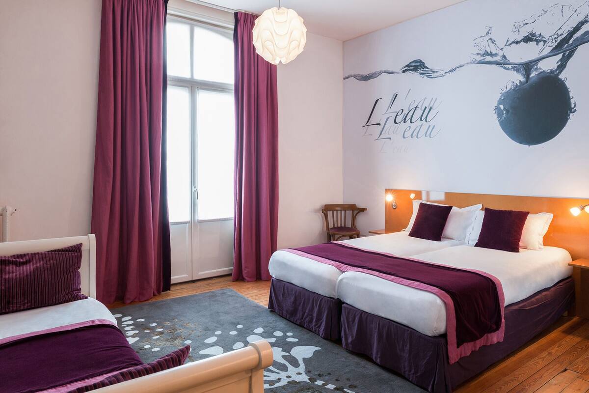 A hotel room with a bed, a chair, a desk, and a large window with curtains.
