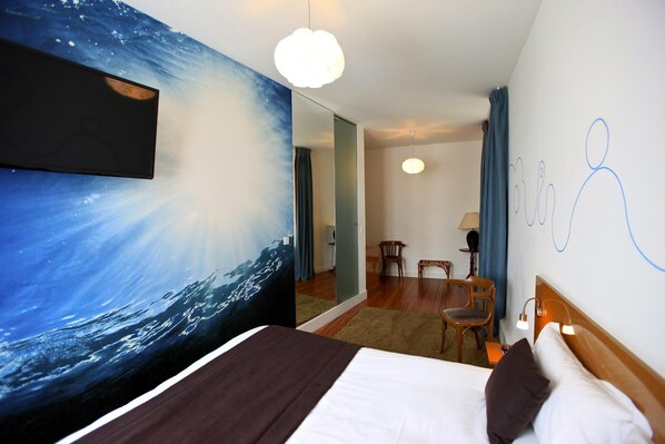 A hotel room with a bed, a large mirror, a painting of the ocean, a wooden chair, and a small table.