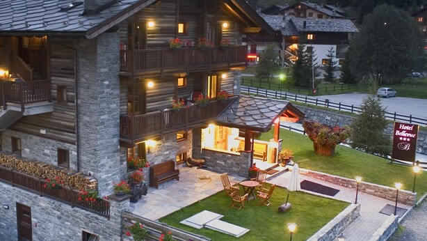 A log cabin hotel with a stone facade, outdoor seating area, and a sign for Hotel Bellevue. - Bellevue (Ayas)