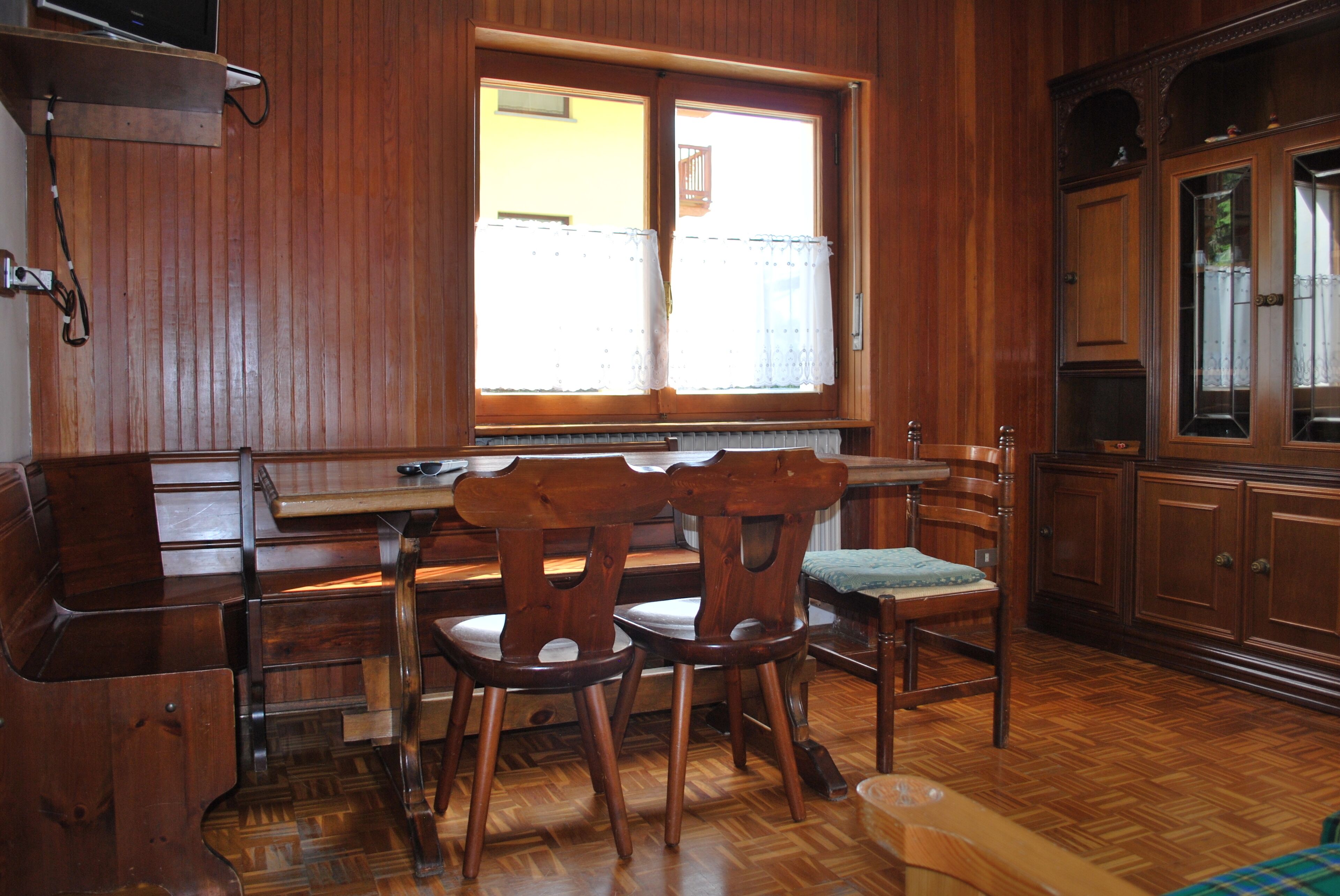 A wooden interior with a dining table and chairs, a window with curtains, and wooden cabinets.