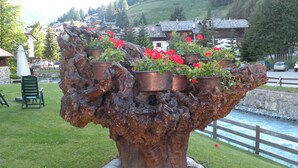A large metal sculpture with potted plants on top, set in a grassy area with a view of houses and a body of water in the background. - Bellevue (Ayas)