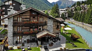 A chalet-style building with a stone facade, wooden balconies, and a riverside patio with outdoor seating. - Bellevue (Ayas)