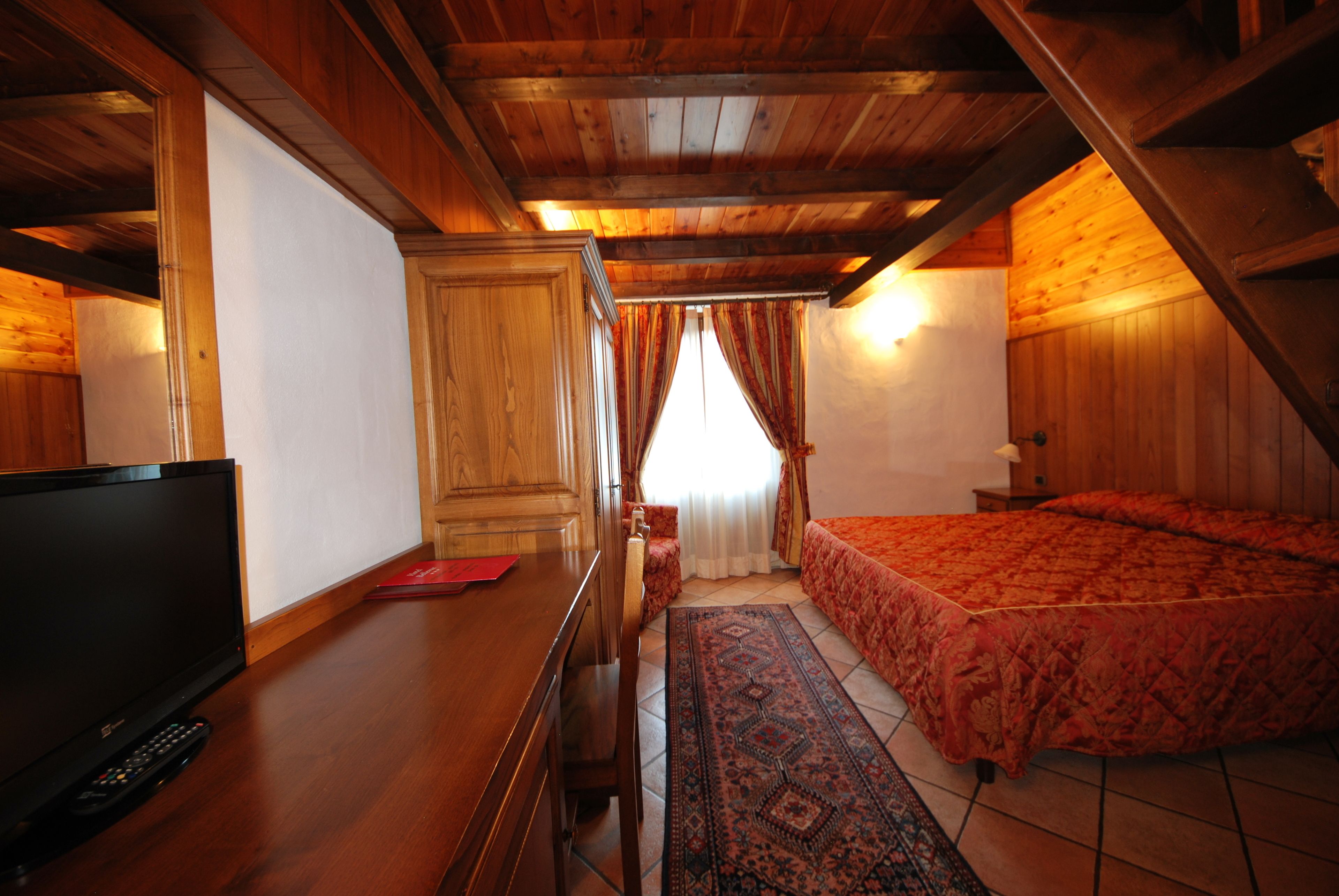 Una habitación con techo de madera, una cama con colcha roja, un televisor sobre una mesa de madera y una alfombra estampada.