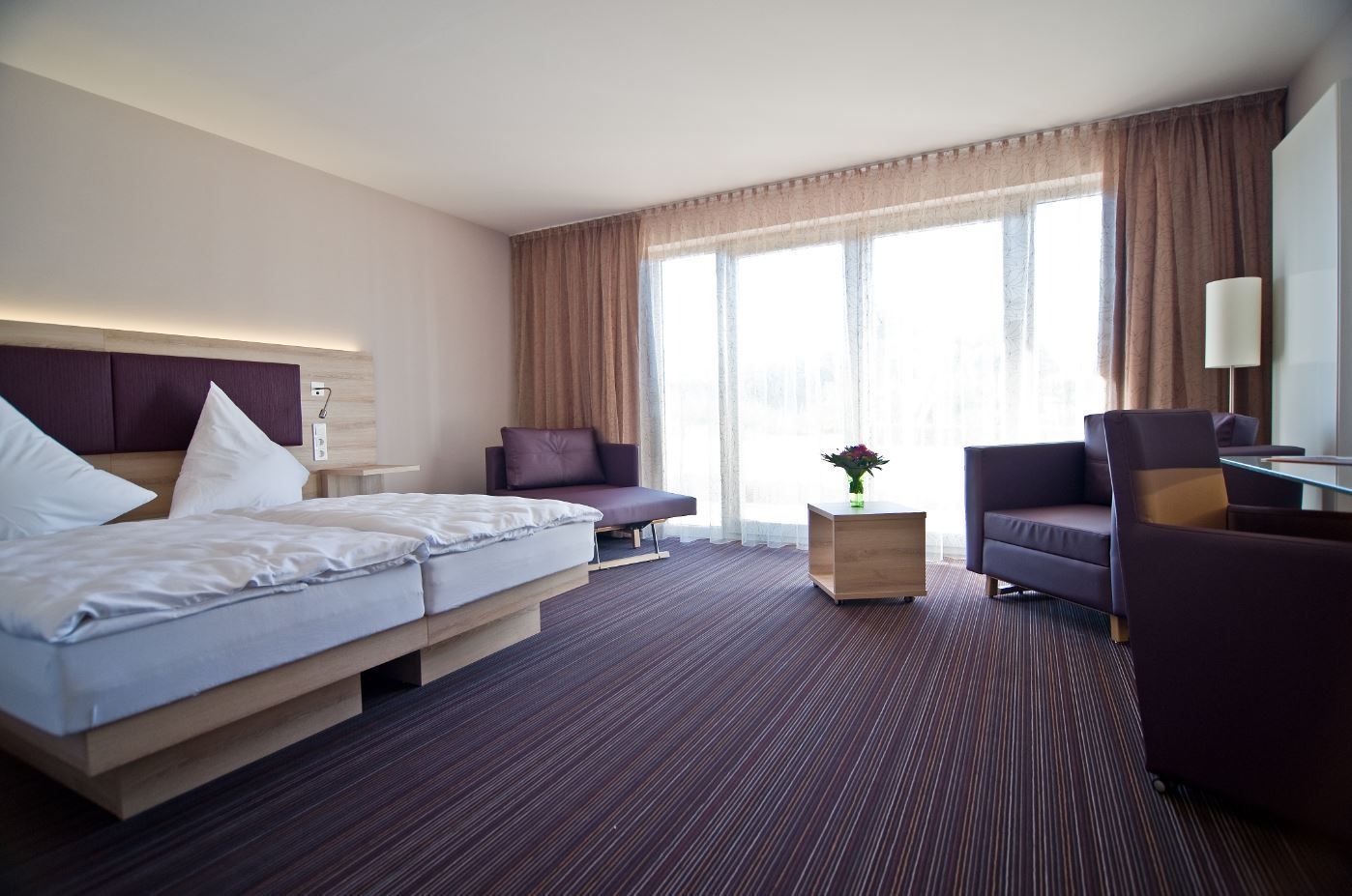Une chambre d’hôtel avec un lit, deux fauteuils, un canapé, une petite table et une grande fenêtre avec des rideaux.