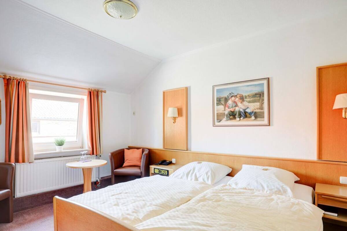 Una camera d'albergo con un letto matrimoniale, un tavolino, una sedia, un comodino, una lampada e una finestra con tende.