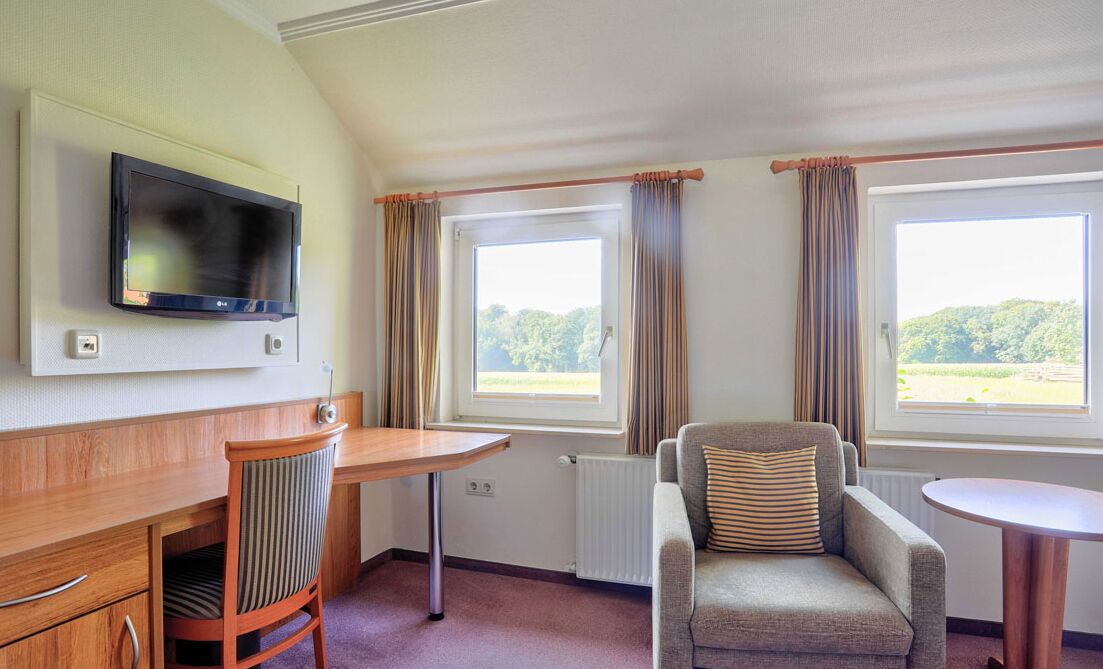 Ein Hotelzimmer mit Schreibtisch aus Holz, einem an der Wand montierten Fernseher, einem gepolsterten Sessel und einem Fenster mit Vorhängen.