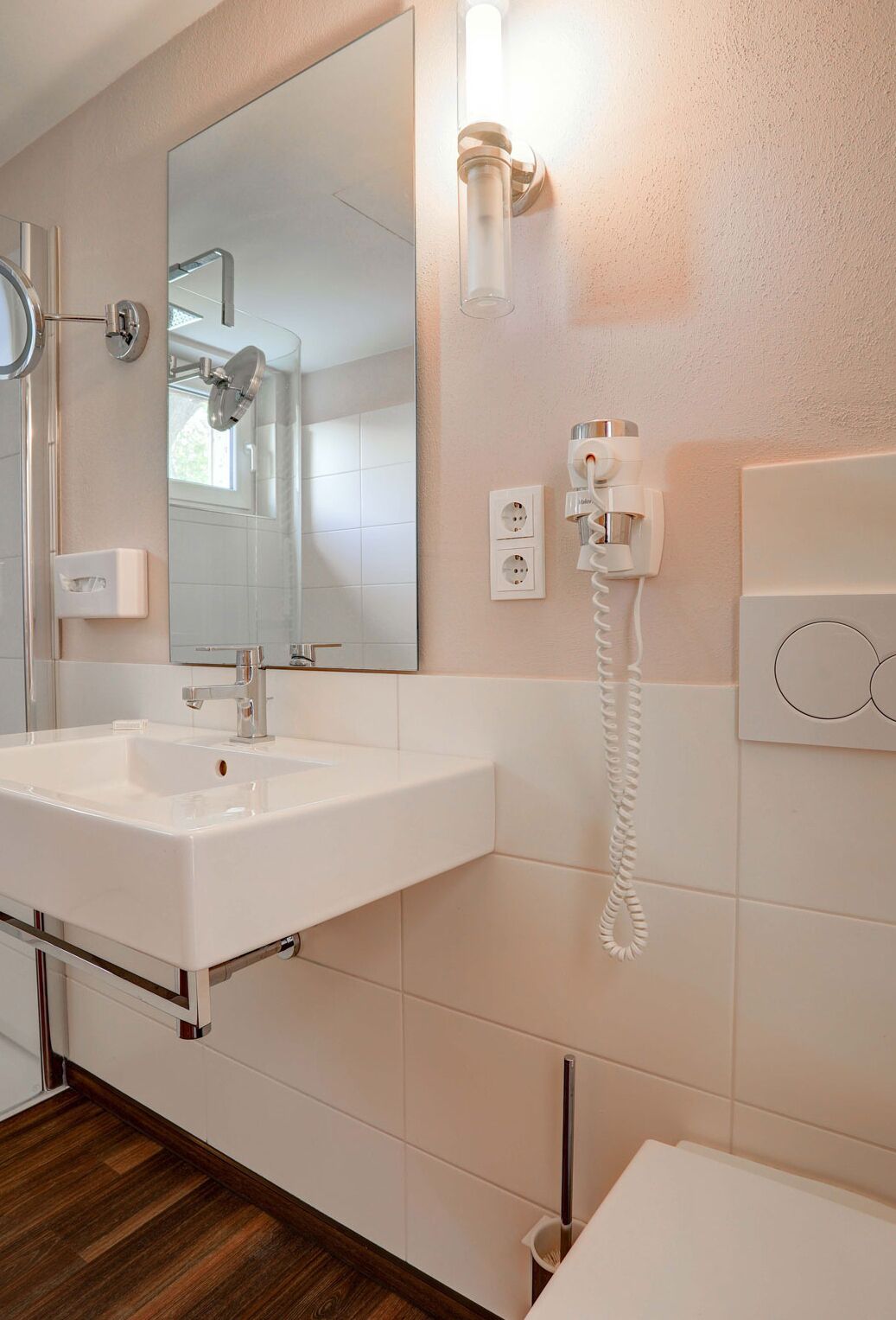 Une salle de bain moderne avec un grand miroir, un lavabo blanc, une douchette à main et un sèche-cheveux fixé au mur.