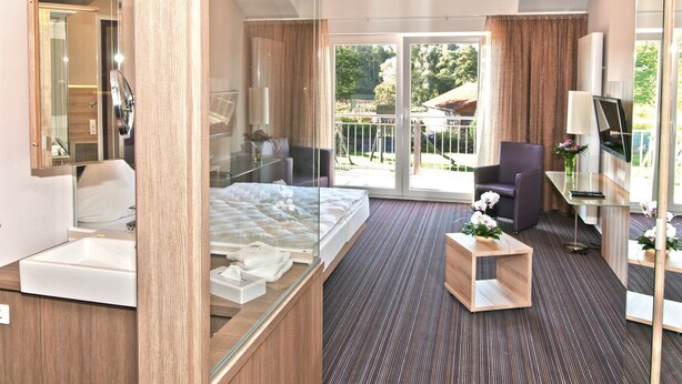 A modern hotel room with a glass-enclosed bed, a desk with a chair, a TV, and a balcony with a view of greenery. - Stössel's Hotel & Restaurant (Altenmedingen)