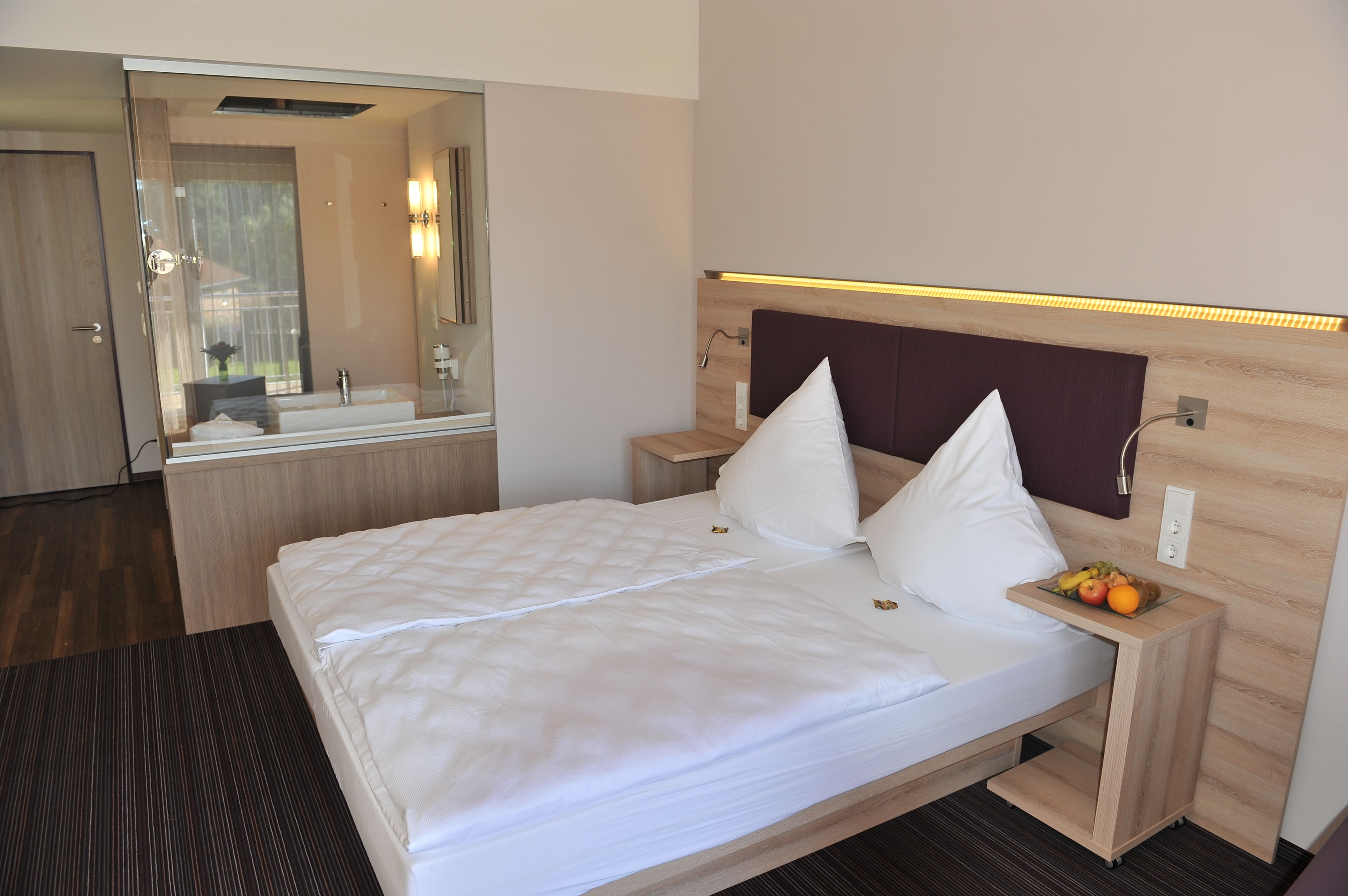 A hotel room with a bed, a nightstand, a mirror, and a small table with fruits.