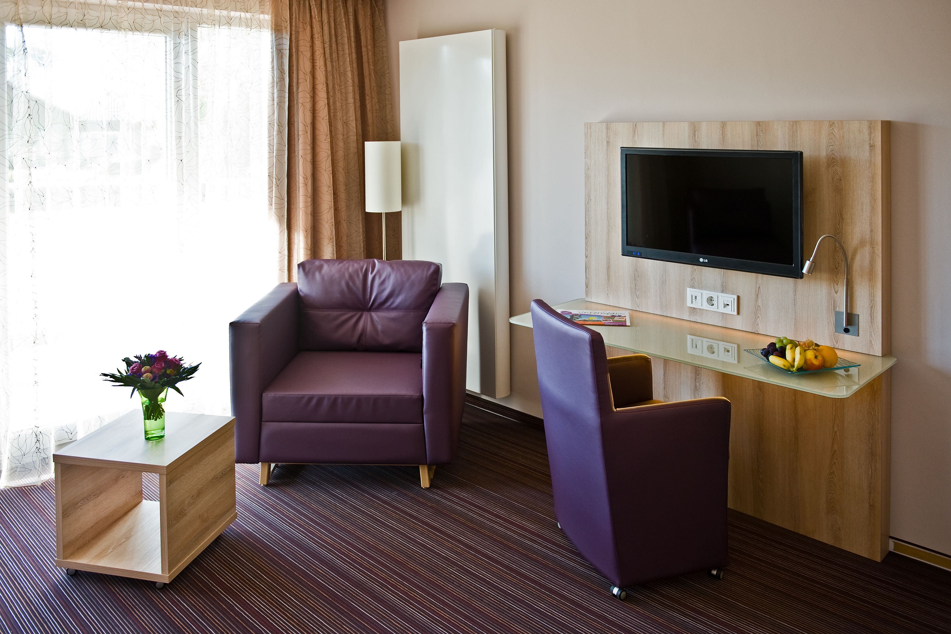 A hotel room with a purple armchair, a small wooden side table with a vase of flowers, a flat-screen TV mounted on a wooden wall, and a glass desk with a fruit bowl.