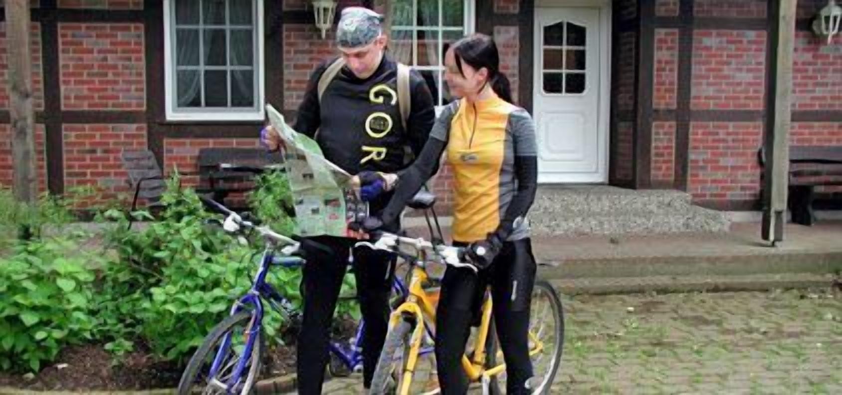 Dos personas de pie junto a bicicletas frente a un edificio de ladrillo con una puerta blanca.