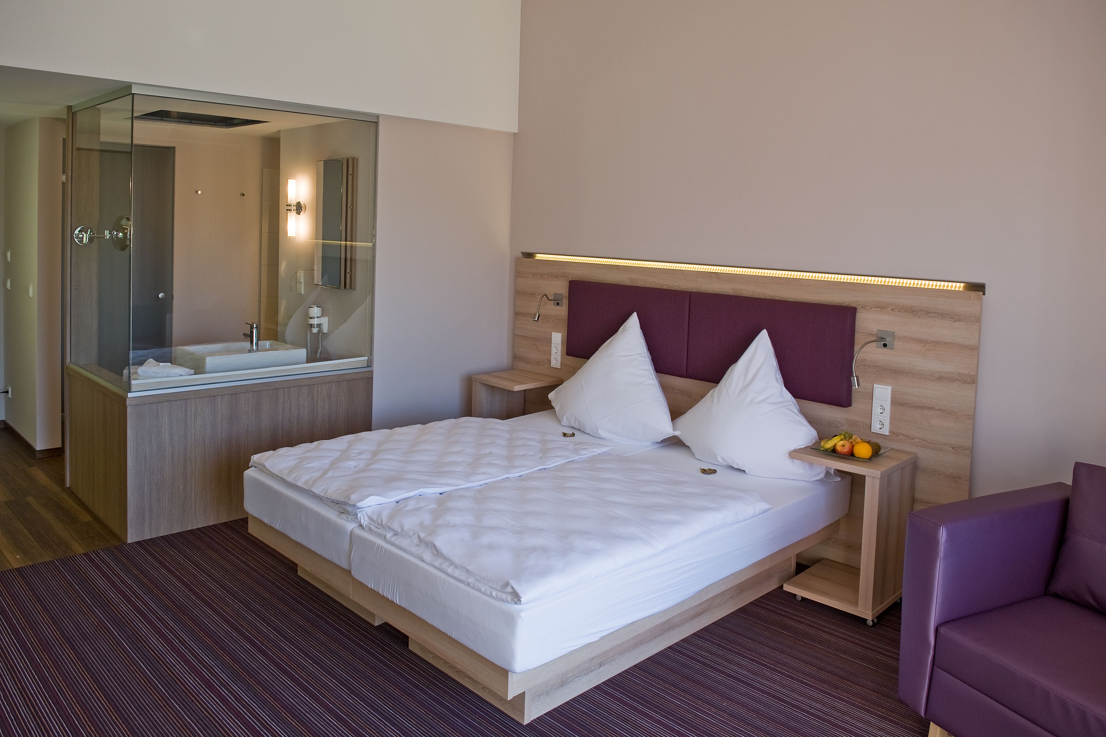A modern hotel room with a large bed, a wooden headboard, a bedside table with a lamp, and a mirror.