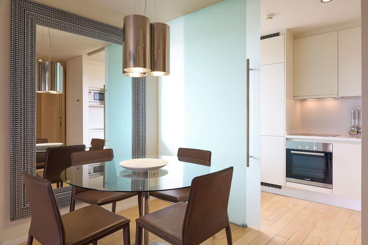 A modern dining area with a glass table and chairs, a large mirror, and a kitchen with white cabinets and a built-in oven.