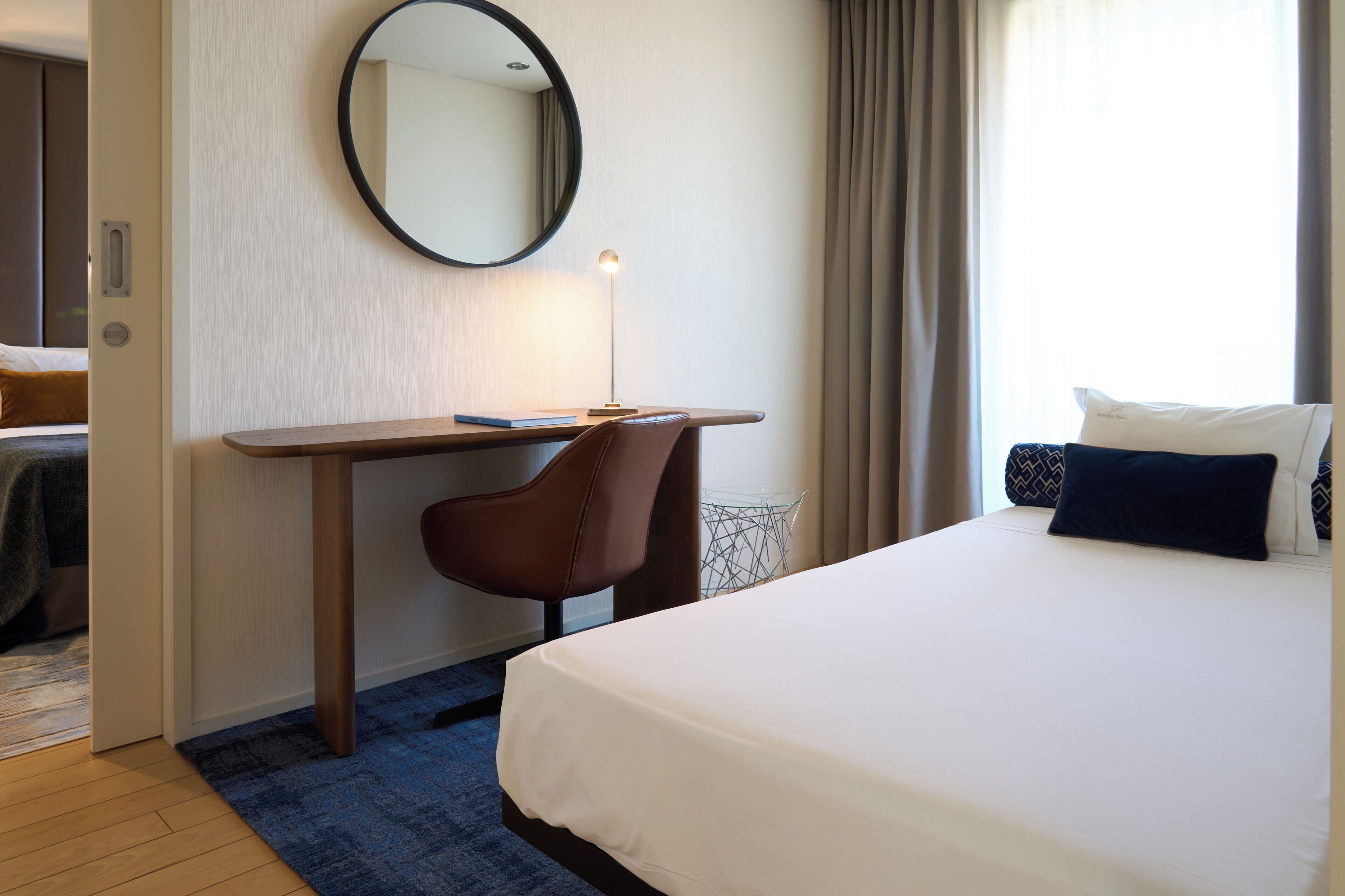 Una habitación de hotel con cama, escritorio y silla, un espejo redondo y una ventana con cortinas.