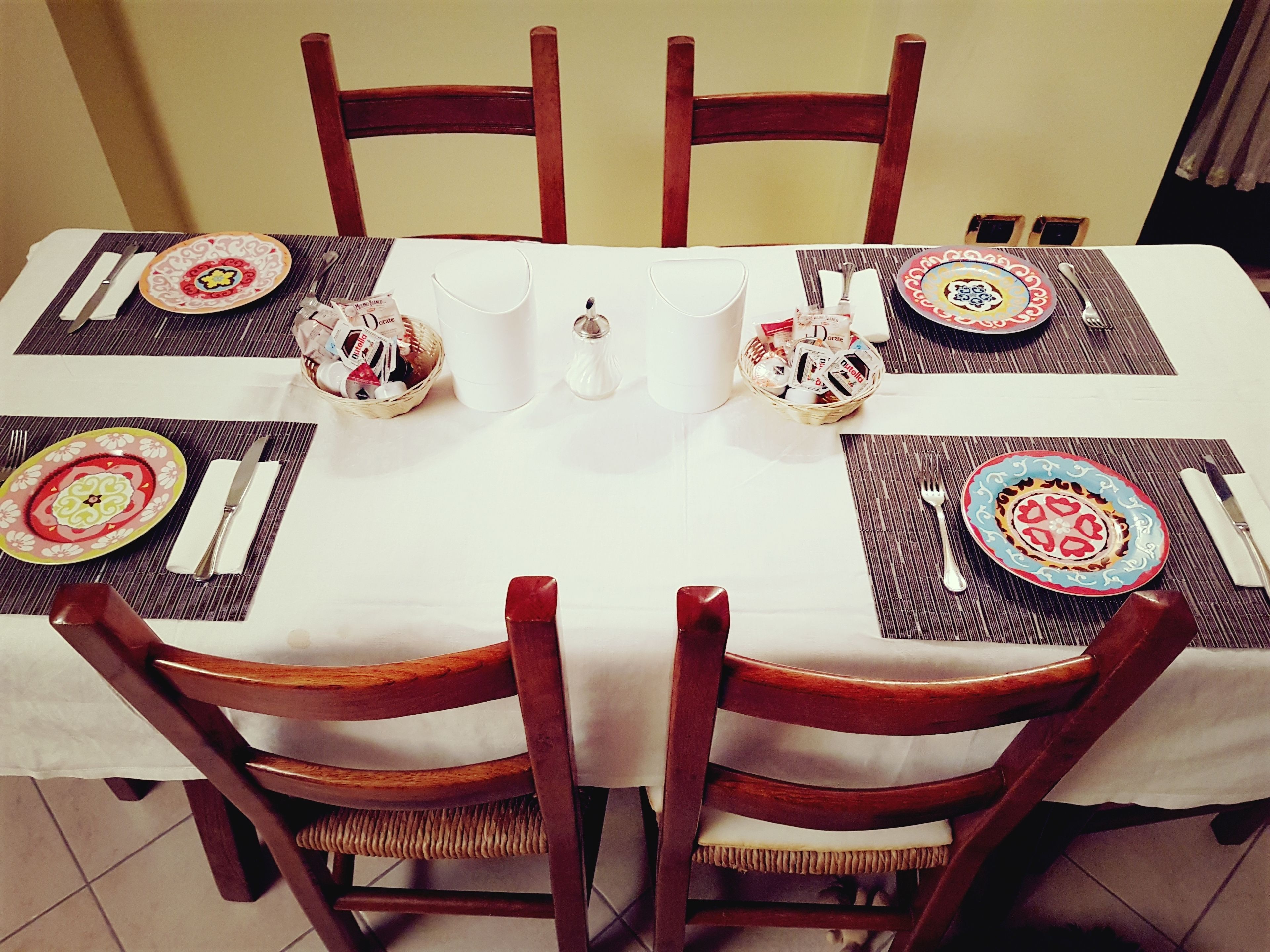 A dining table set for six with a white tablecloth, wooden chairs, and colorful plates.
