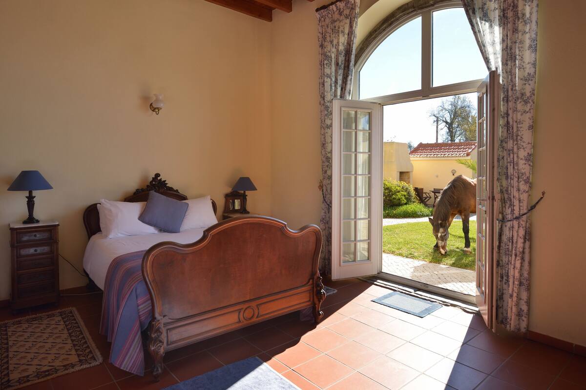 A bedroom with a wooden bed, two bedside tables with lamps, a large window, and a view of a horse outside.