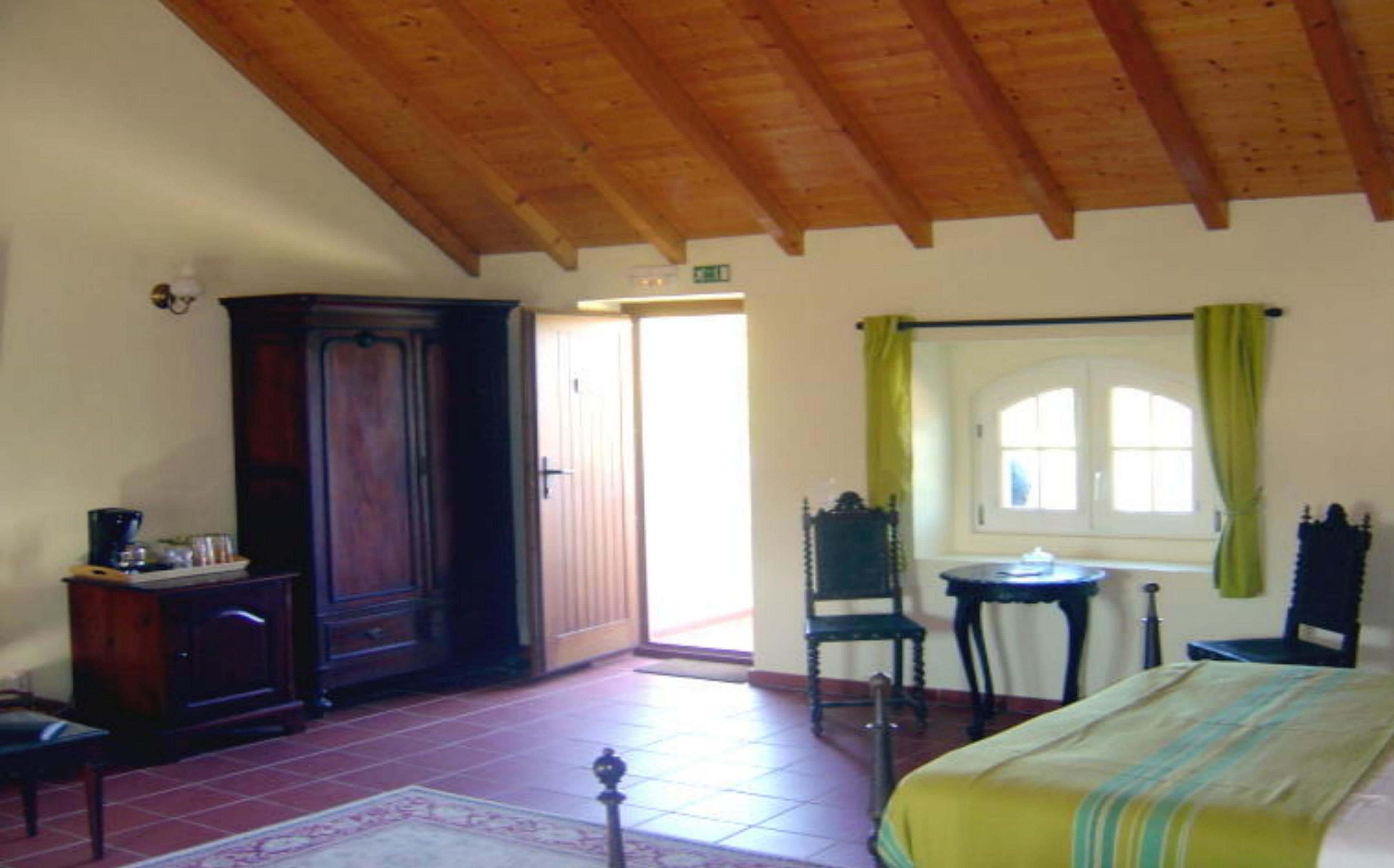 A room with a wooden ceiling, a large wooden wardrobe, a bed with a green and white striped bedspread, a small table with a chair, and a window with green curtains.