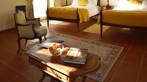 A bedroom with two beds, a ceiling fan, a wooden ceiling, a window with curtains, a chair, a table with a book and a bowl of fruit, and a rug. - Quinta do Scoto (Sintra)