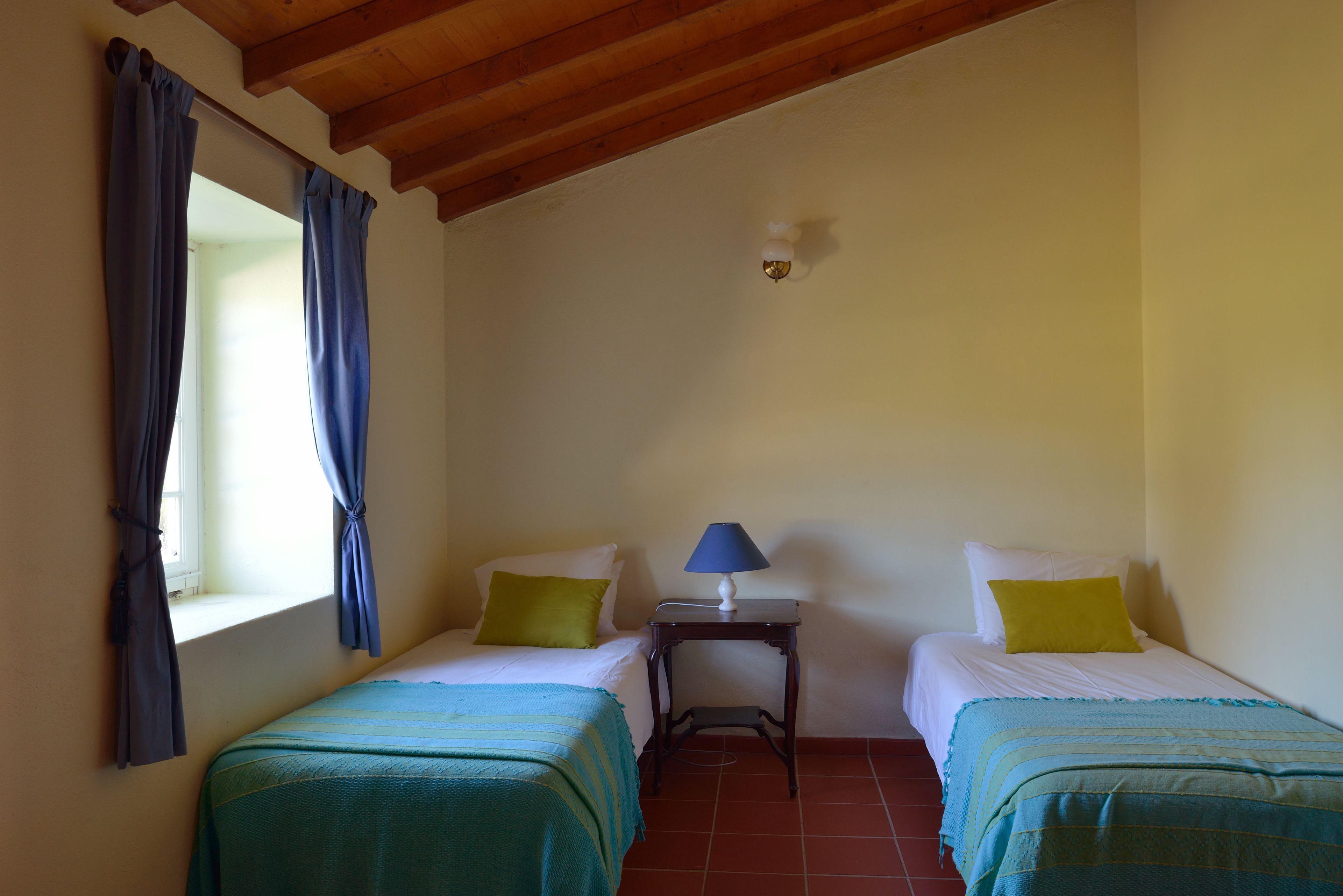 Un dormitorio con dos camas individuales, una mesita de noche con lámpara y una ventana con cortinas.