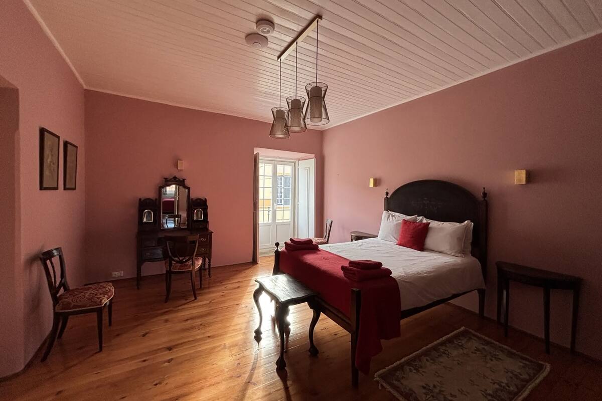 A bedroom with a wooden floor, a bed with red bedding, a desk with a mirror, and a chair.