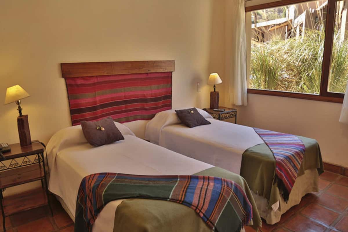A hotel room with two beds, a striped headboard, bedside lamps, a window with a view of greenery, and a tiled floor.