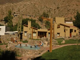 Zona de piscina con muros de piedra y un letrero de madera, rodeada de edificios de estilo desértico.