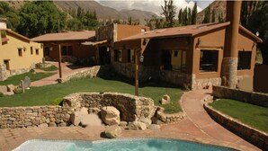 A pool area with a stone border, a stone wall, and a building with a brown roof. - Hotel El Reposo del Diablo (Tilcara)