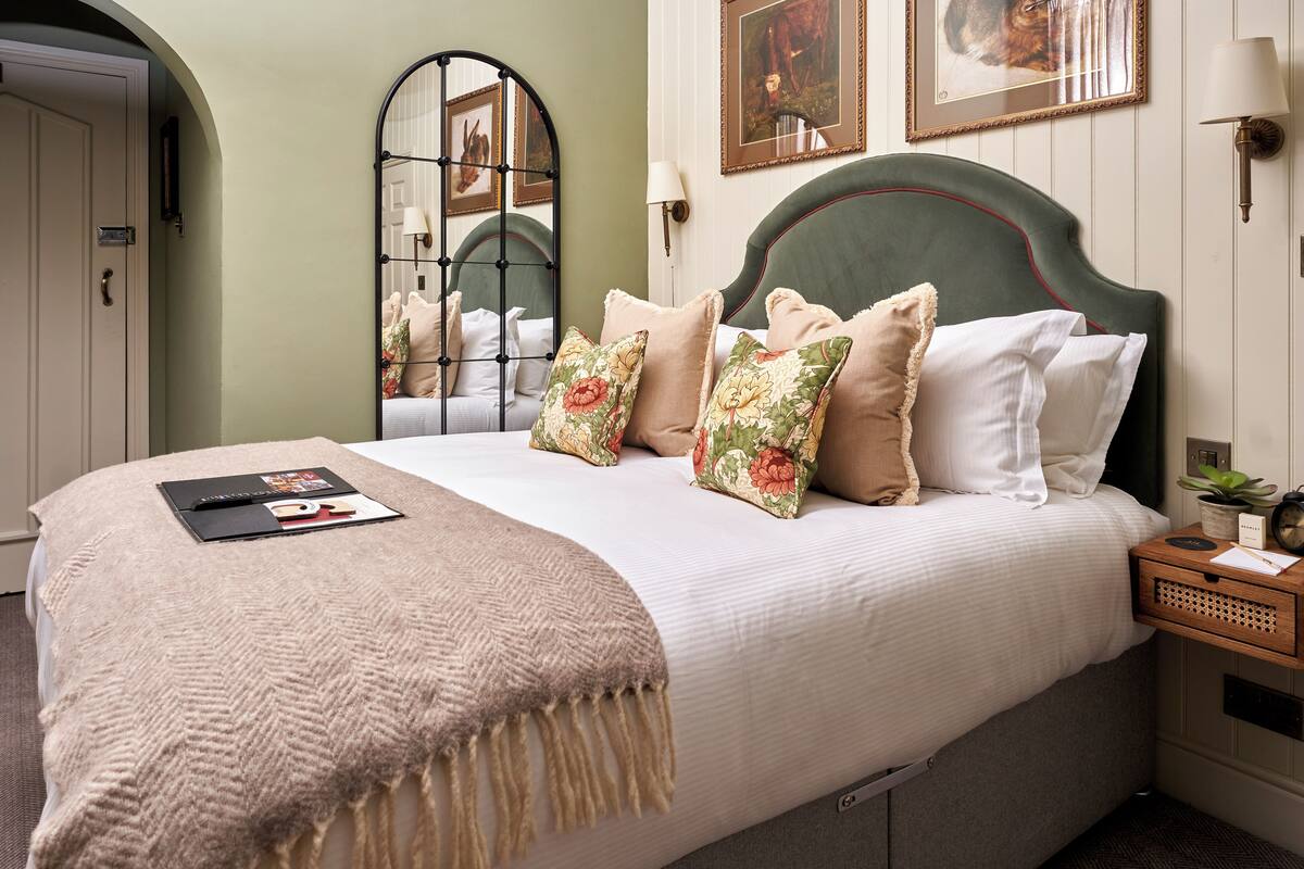 A neatly made bed with a beige blanket, floral pillows, and a small bedside table with a lamp and a plant.