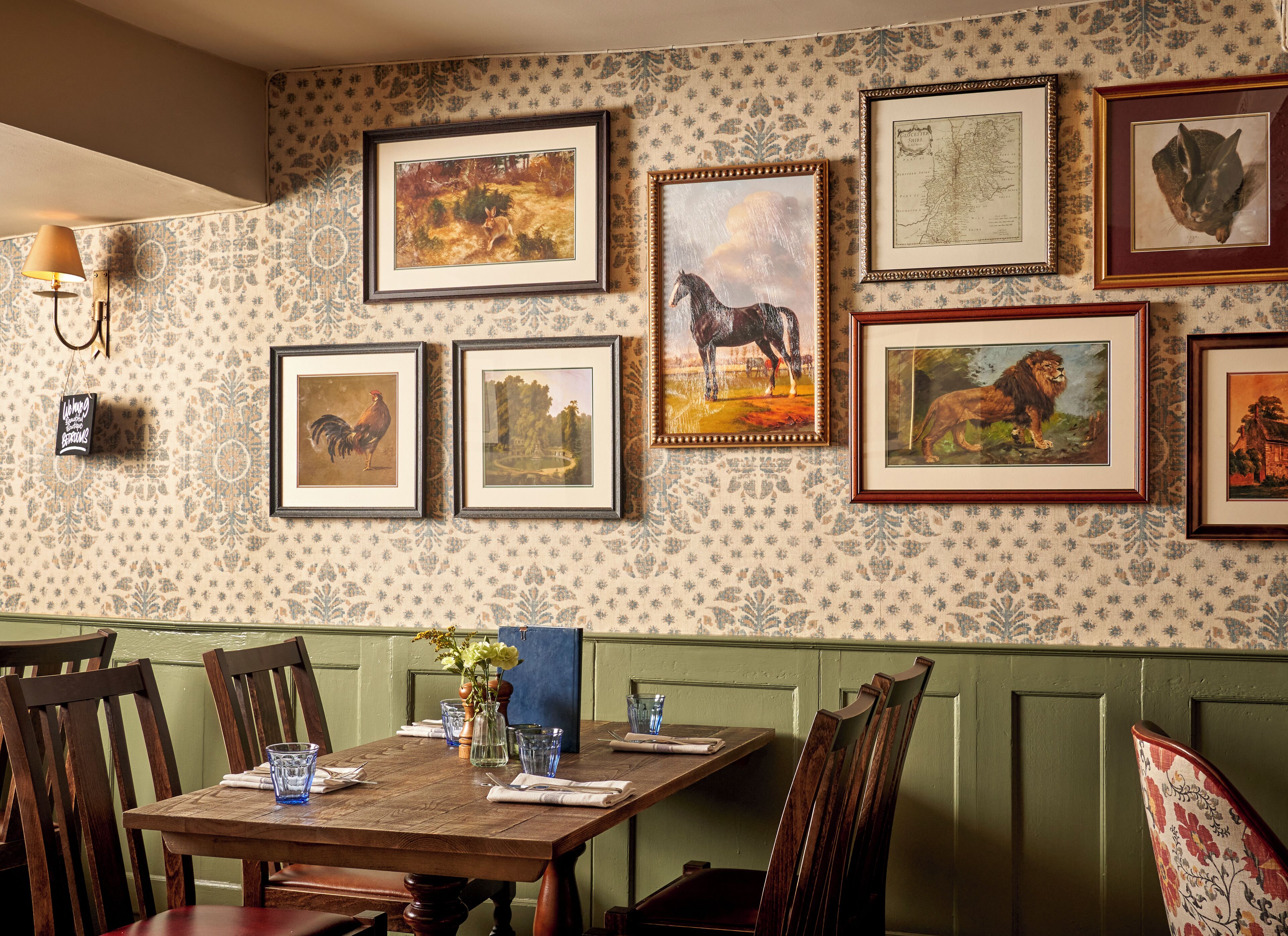 Un coin repas avec une table en bois dressée pour quatre personnes, entourée de chaises en bois. Le mur est décoré d’œuvres d’art encadrées représentant des animaux et des paysages.