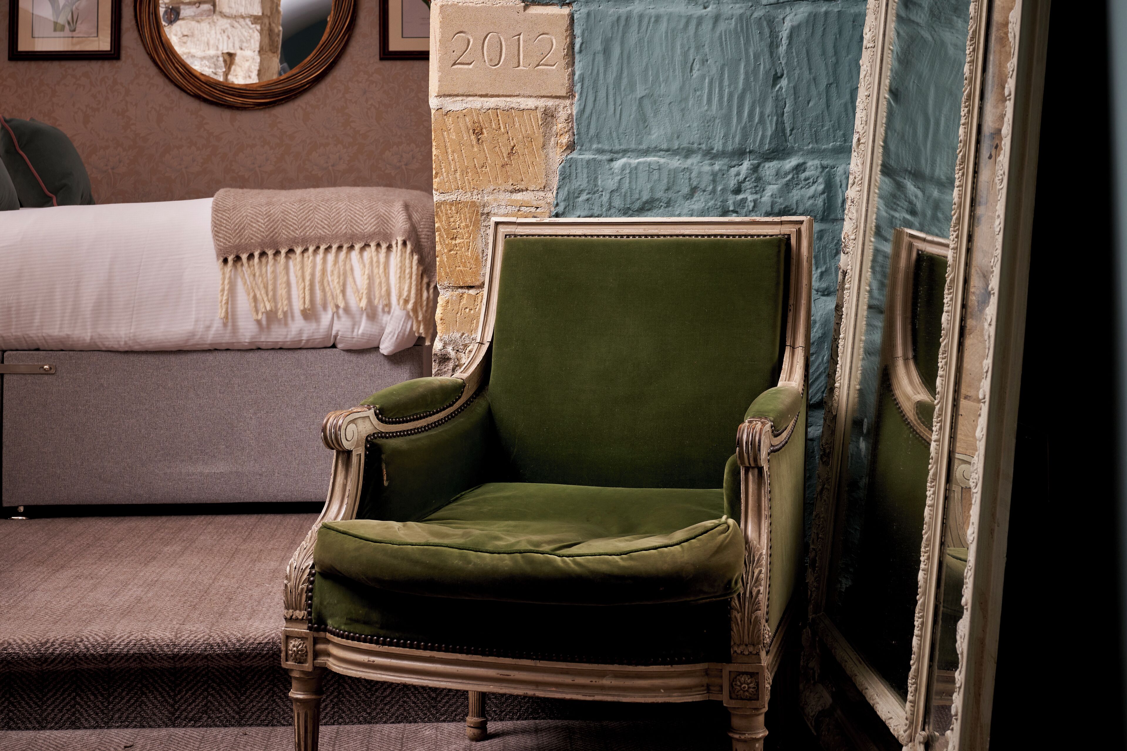 Un sillón tapizado de verde en una habitación con pared de ladrillo, una cama con colcha blanca y un espejo con marco decorativo.