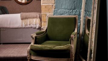 Un sillón tapizado de verde en una habitación con pared de ladrillo, una cama con colcha blanca y un espejo con marco decorativo.