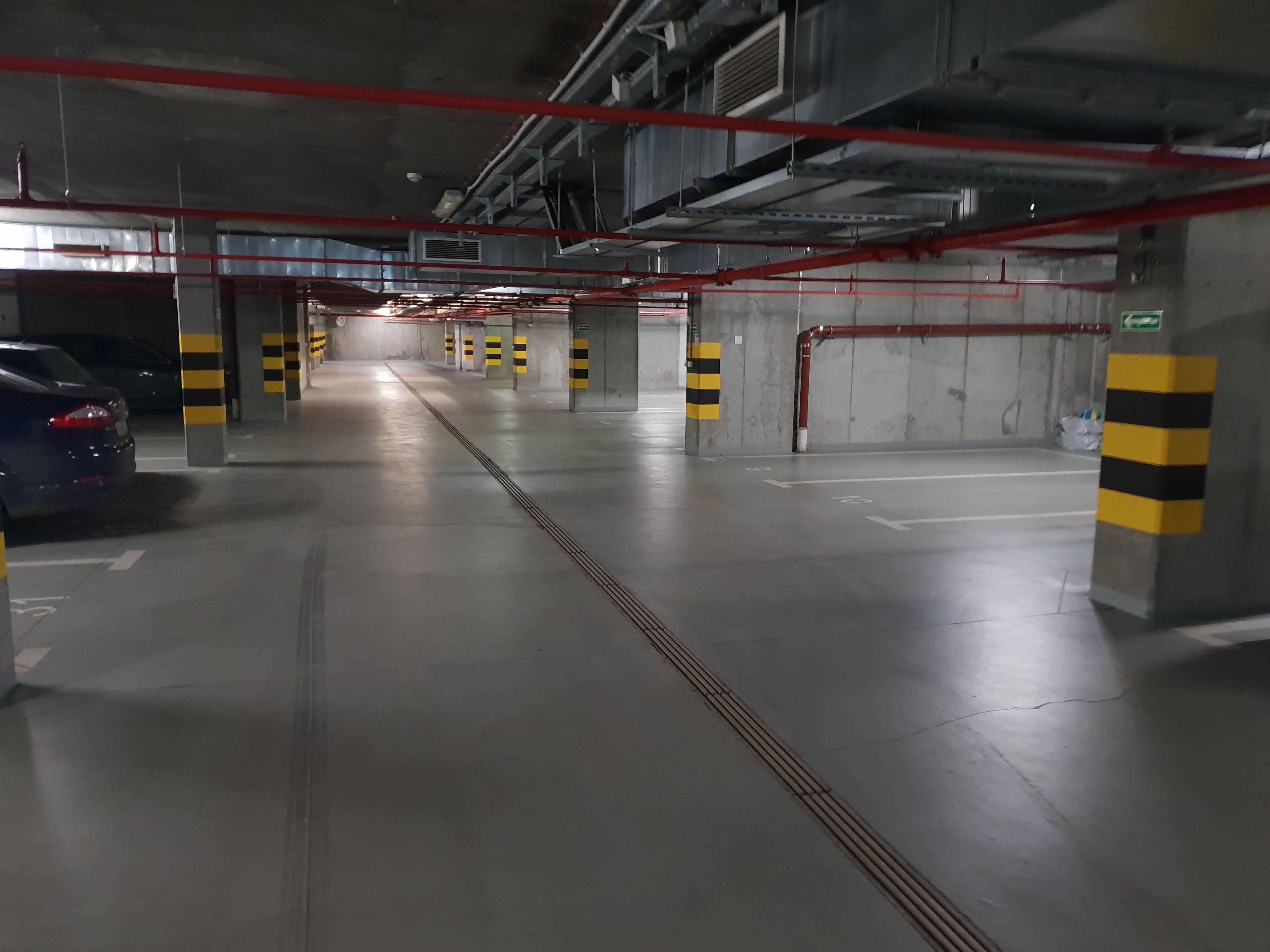An underground parking garage with concrete pillars, yellow and black striped bollards, and red pipes on the ceiling.