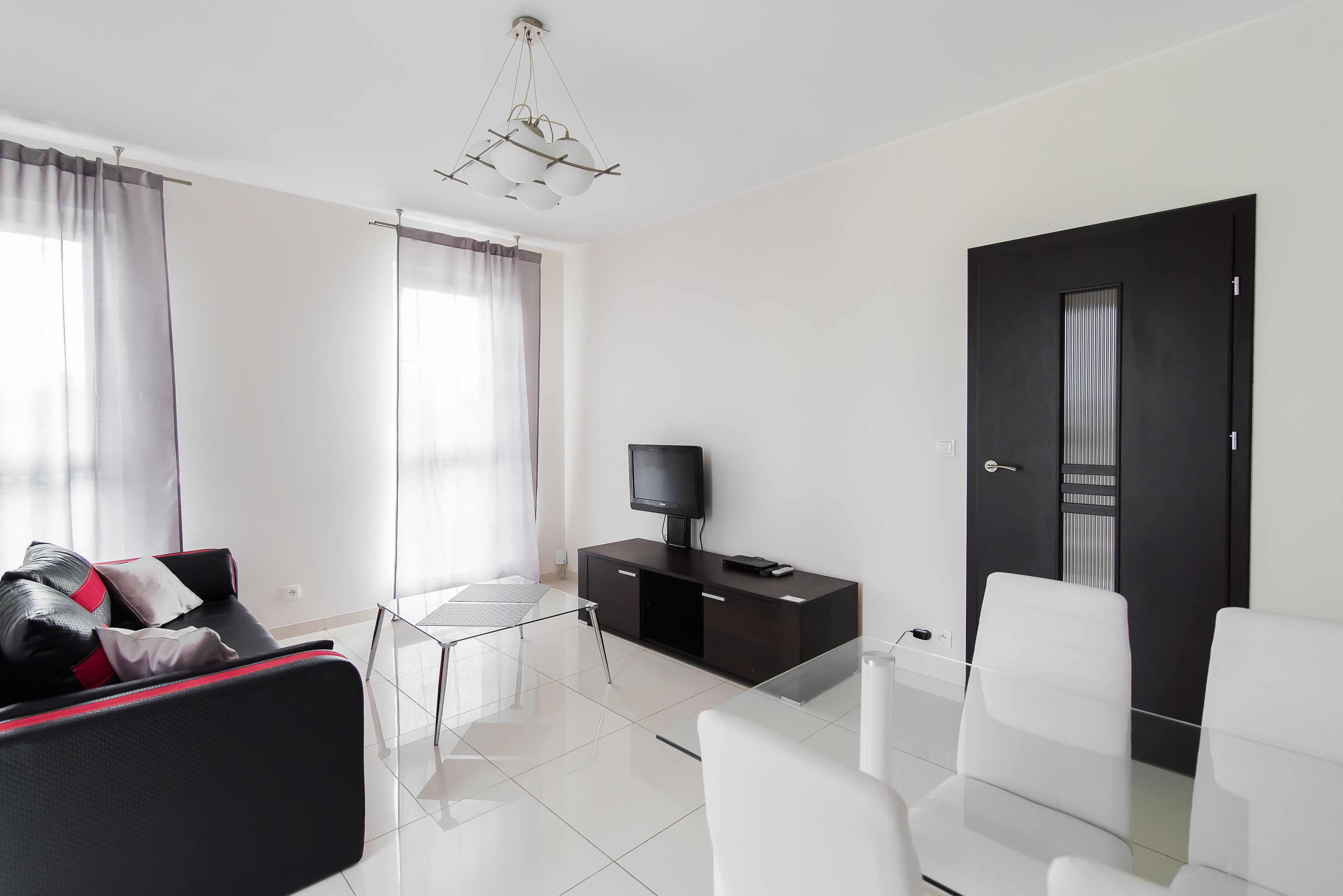 Una camera d'albergo moderna con un divano nero, un tavolino in vetro, sedie bianche, una TV su un mobile nero e una porta con una striscia verticale.