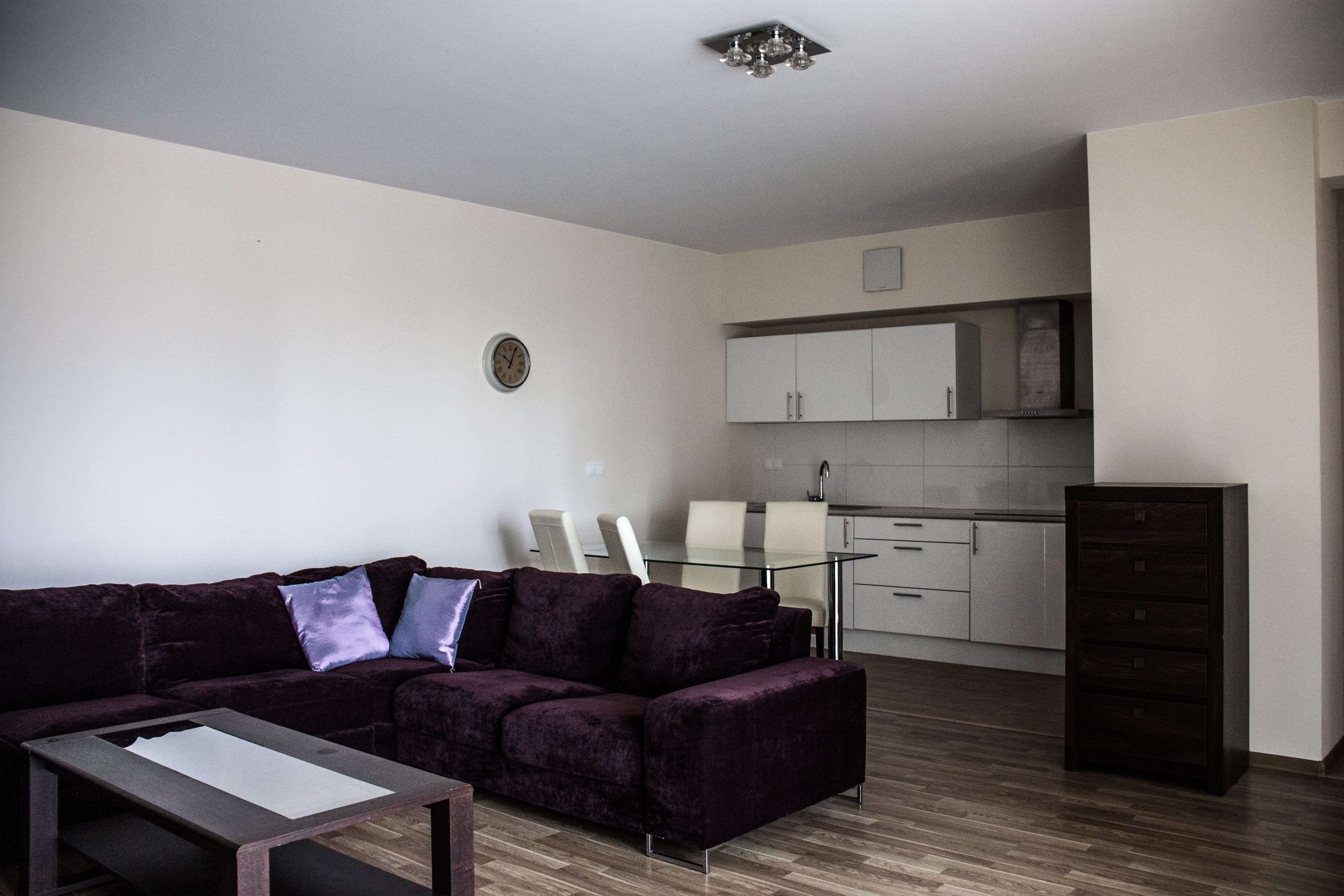 Un salon moderne avec un canapé violet foncé, une table basse en verre et un coin repas comprenant une table et des chaises en verre. On aperçoit, au fond, une cuisine avec des meubles blancs et un meuble en bois foncé.