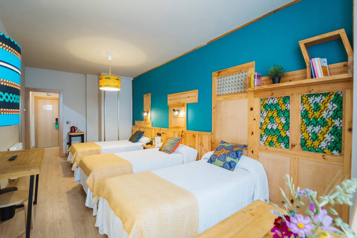 Quarto de hotel com duas camas de solteiro, uma escrivaninha de madeira, uma mesinha, uma cadeira, um espelho, uma estante com livros e uma planta, e uma decoração de parede colorida.
