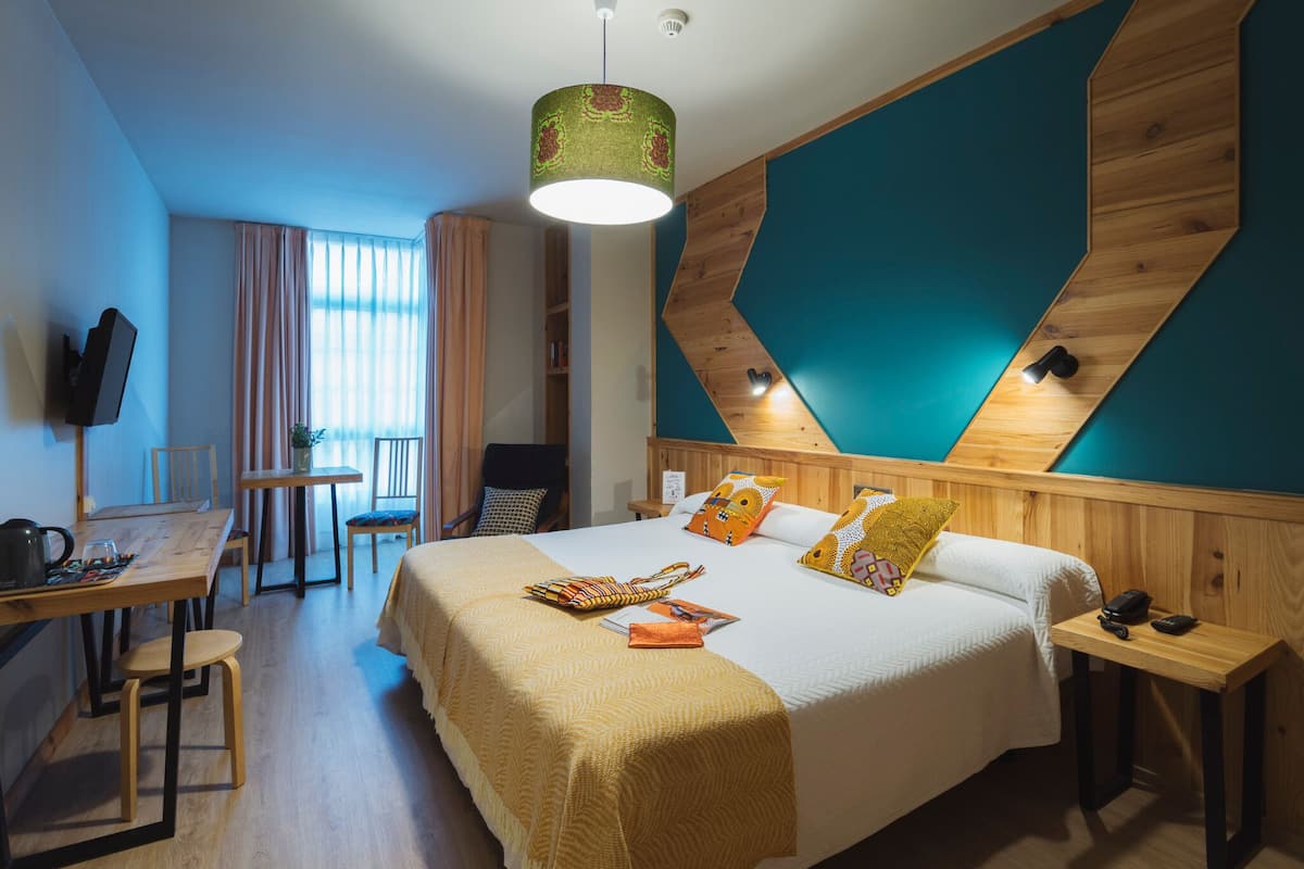 Quarto de hotel com cama, escrivaninha, cadeira, televisão e janela com cortinas.