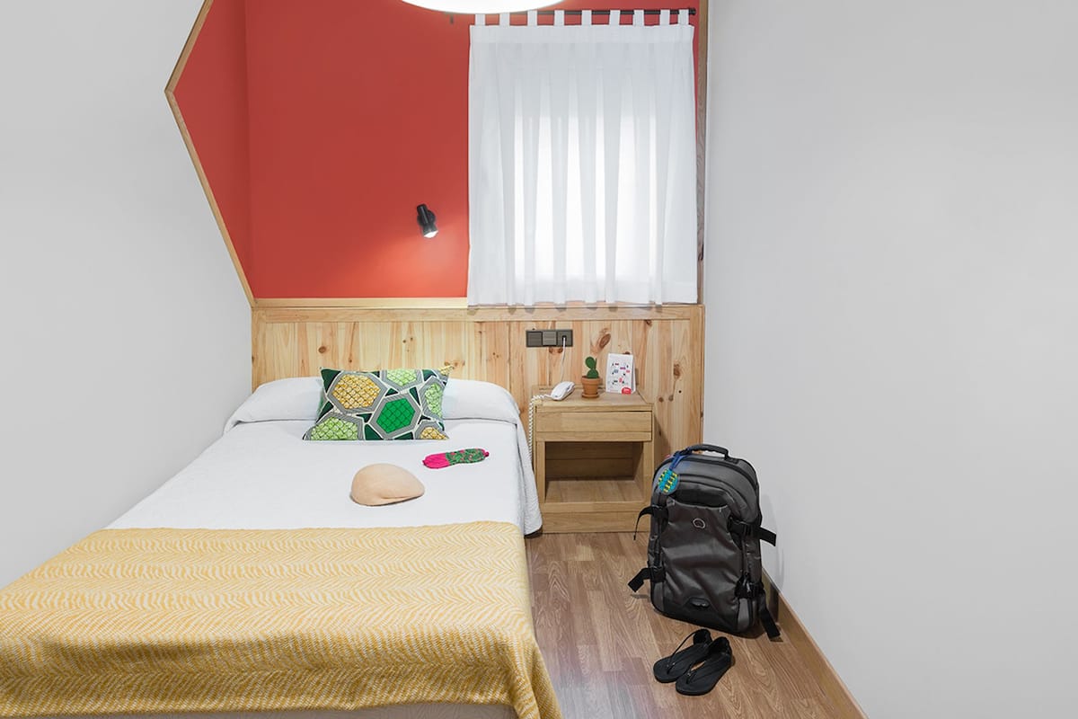 Quarto com cama, mesa de cabeceira, cabeceira de madeira, janela com cortinas brancas e uma mochila no chão.