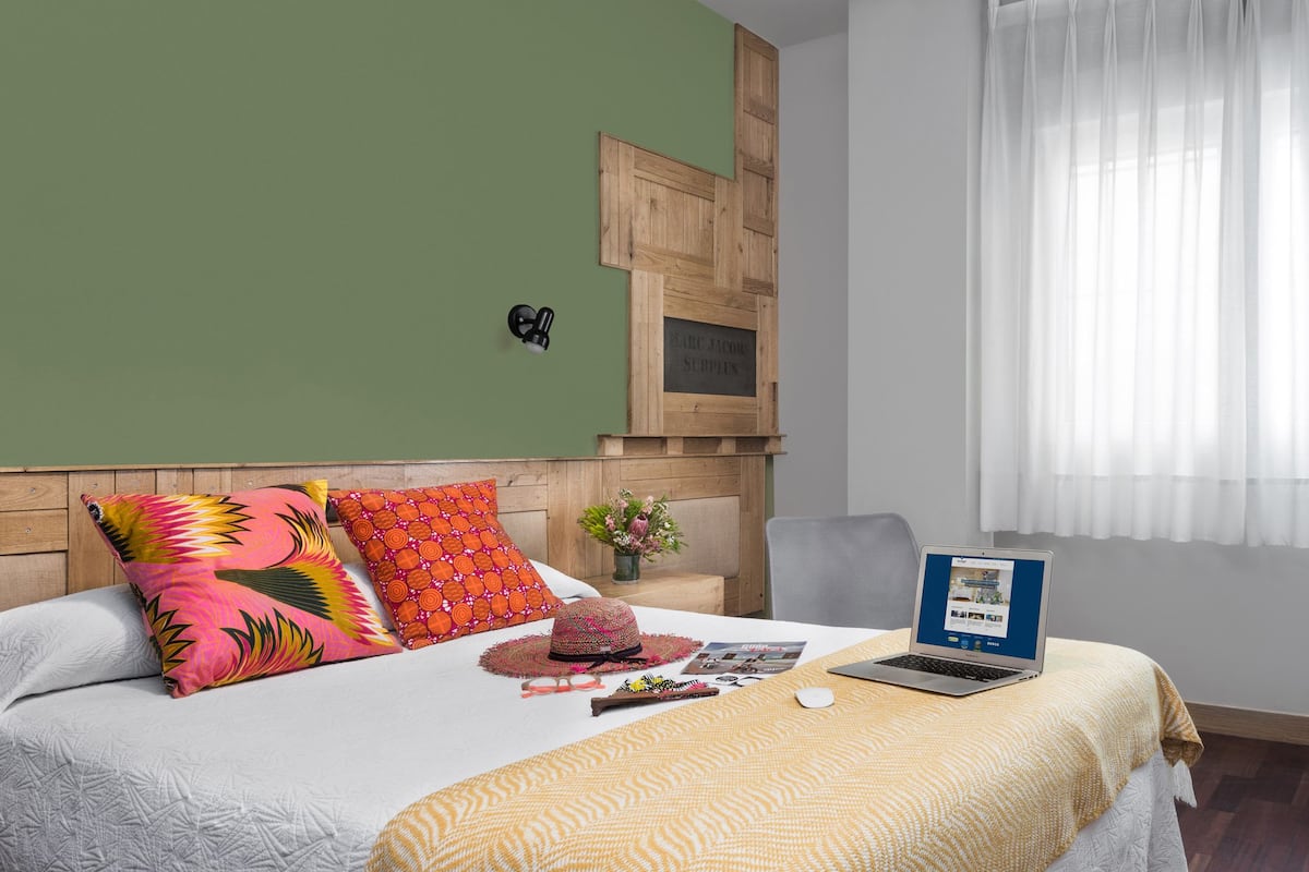 Quarto de hotel com cama, um laptop, escrivaninha com abajur e janela com cortinas.