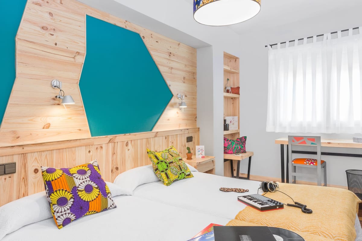 Quarto de hotel moderno com cabeceira de madeira, cama com almofadas coloridas, escrivaninha com cadeira e janela com cortinas brancas.