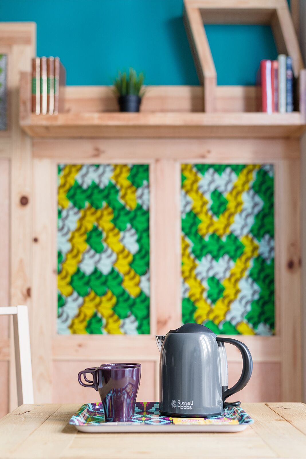 Una mesa de madera con una taza y una tetera, un mantel individual colorido y una estantería con libros y una pequeña planta.
