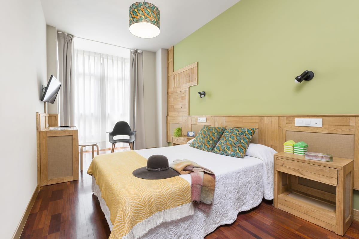 Quarto de hotel com cama de madeira, uma escrivaninha com luminária, uma cadeira, mesa de cabeceira com luminária e uma janela com cortinas.