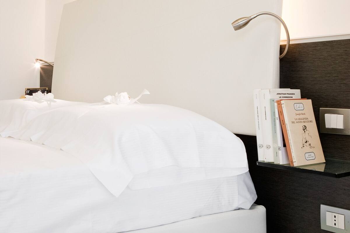 Ein ordentlich bezogenes Bett mit weißen Leinen, ein Nachttisch mit Lampe und einige Bücher im Regal.