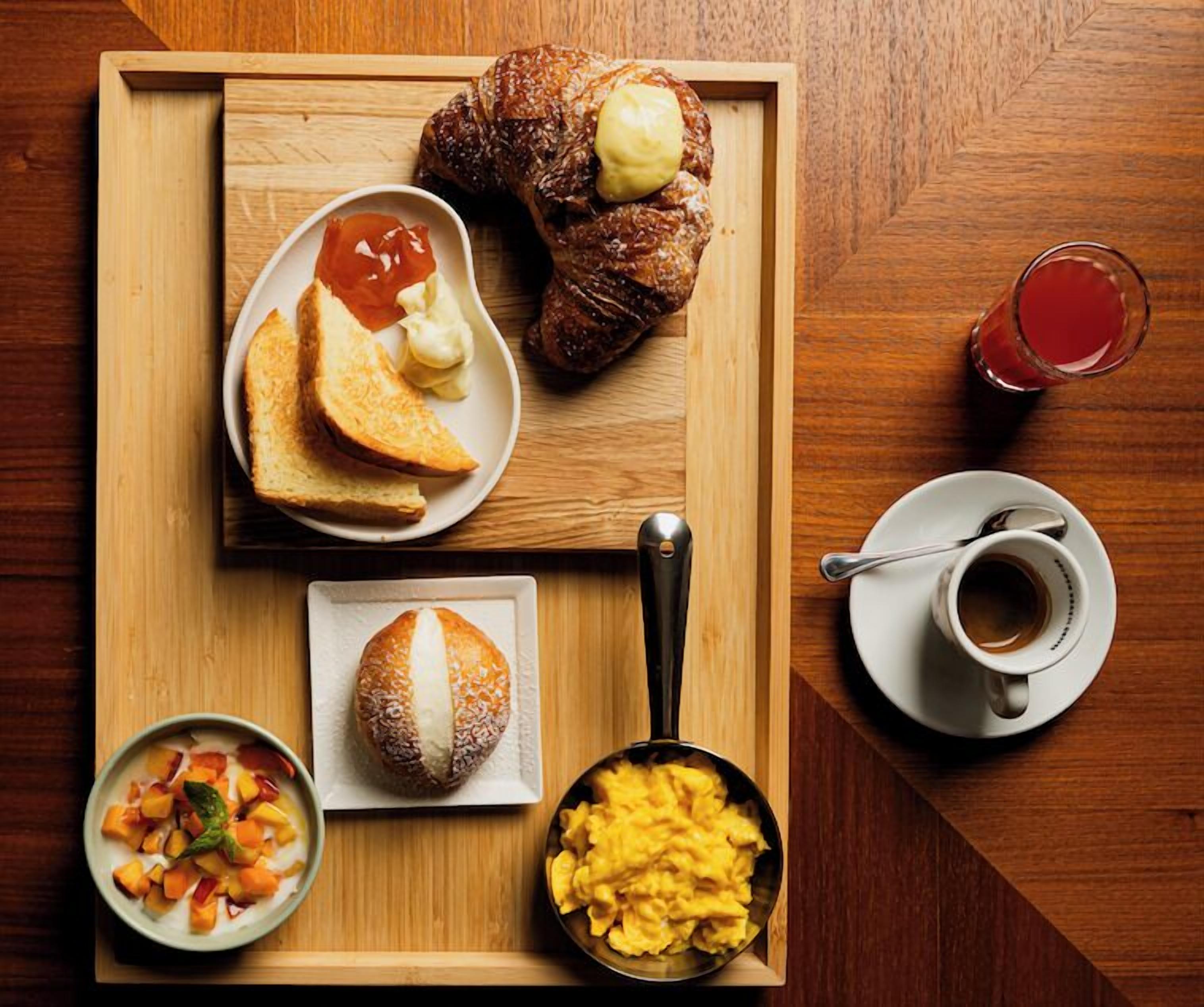 Ein Holztablett mit Croissants, Toast, Rührei und einer Schüssel Obstsalat.