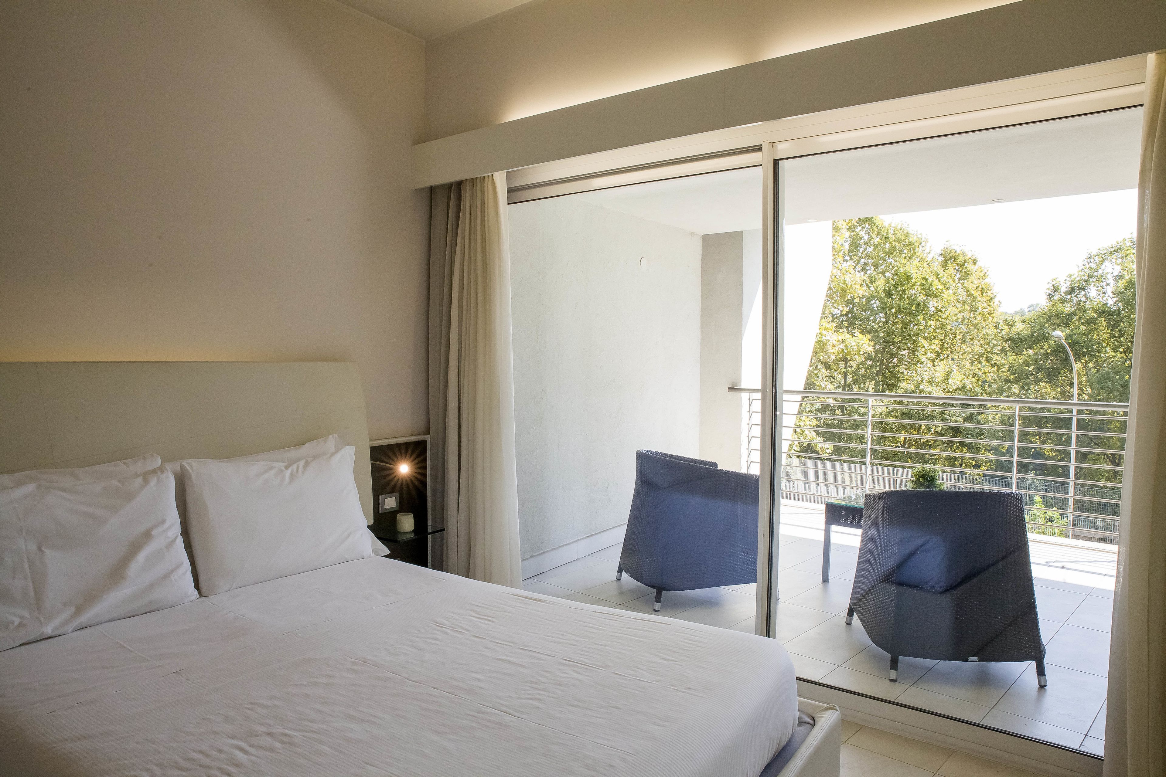 A hotel room with a large bed, a balcony with two chairs, and a view of greenery outside.