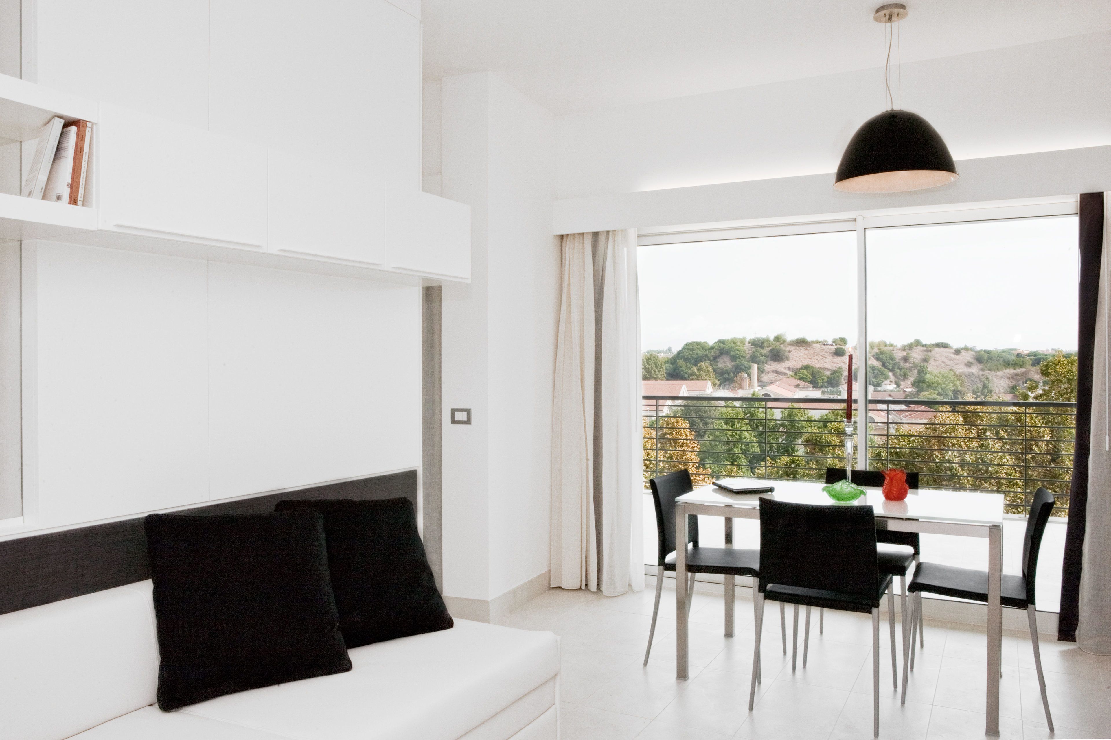 Una sala de estar moderna con un sofá blanco, cojines negros, una mesa de comedor con sillas y un ventanal que ofrece vistas a un paisaje montañoso.