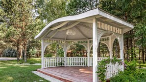 Un pabellón con terraza de madera y barandillas blancas, rodeado de follaje y árboles.