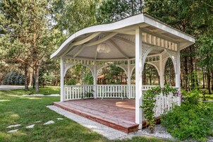 Um gazebo com deck de madeira e grades brancas, rodeado por vegetação e árvores.
