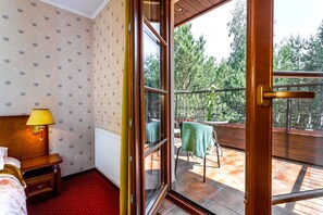 A hotel room with a wooden headboard, a bedside lamp, a window with a view of the outdoors, and a balcony with a table and chairs. - Hotel Dworek Tryumf Dąbrowscy Spółka Jawna (gmina Juchnowiec Koscielny)